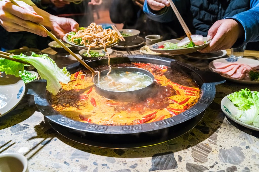 A divided Sichuan hotpot on the table, chopsticks lifting food out of the chilli broth