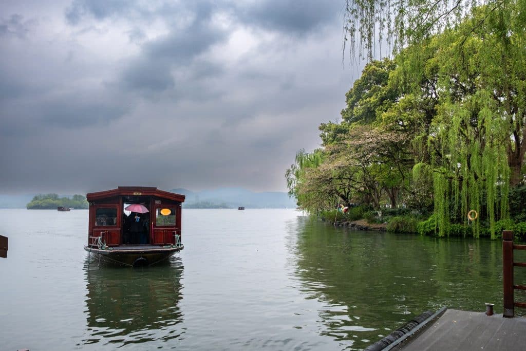 A traditional wooden tour boat on West Lake in Hangzhou, with weeping willow trees along the shore and misty hills in the distance