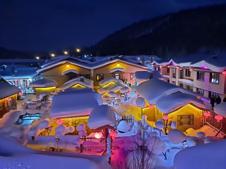 China Snow Town at night, wooden houses with thick rounded caps of snow on their roofs, lit by red lanterns and coloured lights
