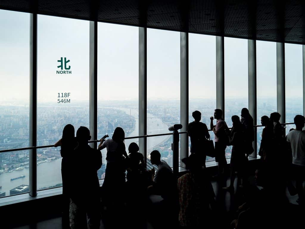 Visitors silhouetted against the floor-to-ceiling windows of the Shanghai Tower observation deck, with a north marker on the glass